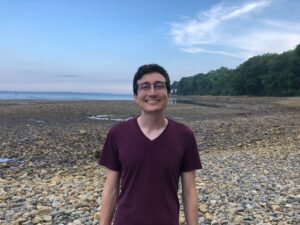 Isaac Goldstein stands on a rocky beach with trees in the background. He is smiling, with dark hair, glasses, and fair skin. He is wearing a maroon v-neck t-shirt.