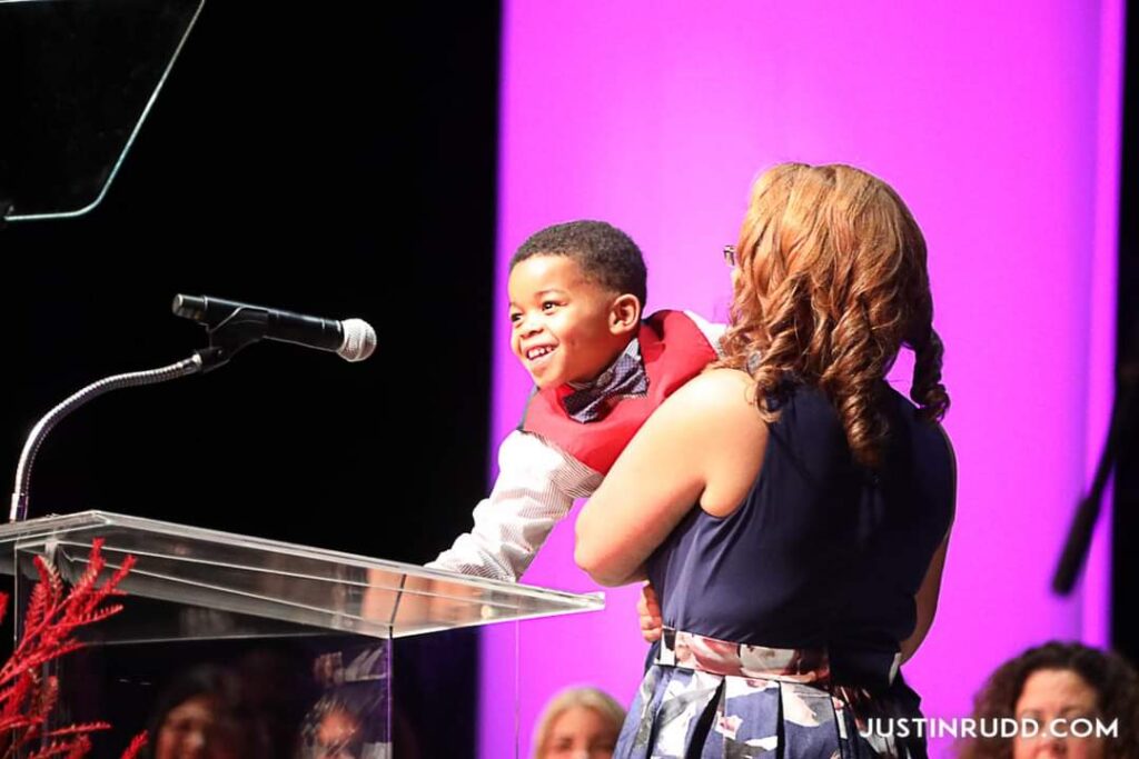 Young boy in mother’s arms, leaning toward a microphone.