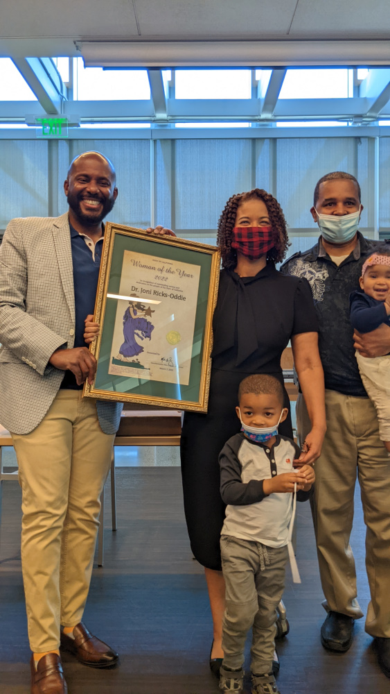 Joni is standing, holding one side of a framed “Woman of the Year” award. Her husband is by her side, holding their young daughter, and her son is standing in front of her. Assembly member Mike Gipson is standing, holding the other side of the framed award.