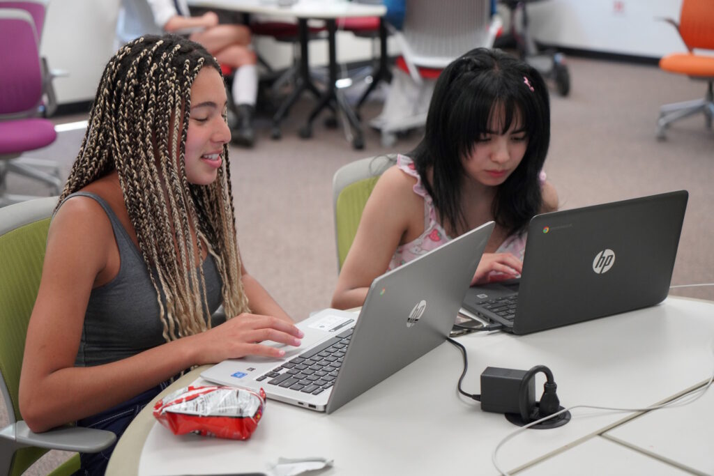 Two students sitting next to each other, each working on a laptop.