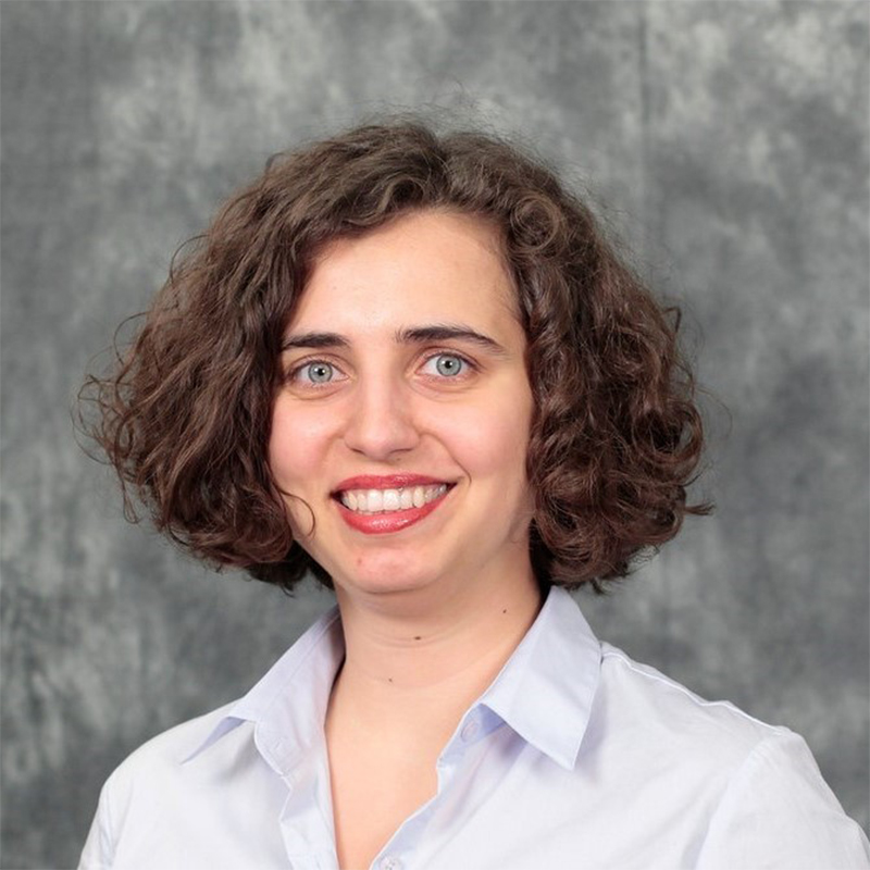 A headshot of Mine Dogucu; Dogucu has chin-length, thick, wavy brown hair and big, light blue eyes. She is smiling broadly and wearing a white blouse with a gray backdrop.